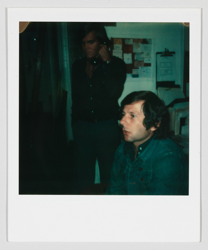 Two men in an office; one is sitting and looking ahead, the other is standing and talking on the phone.