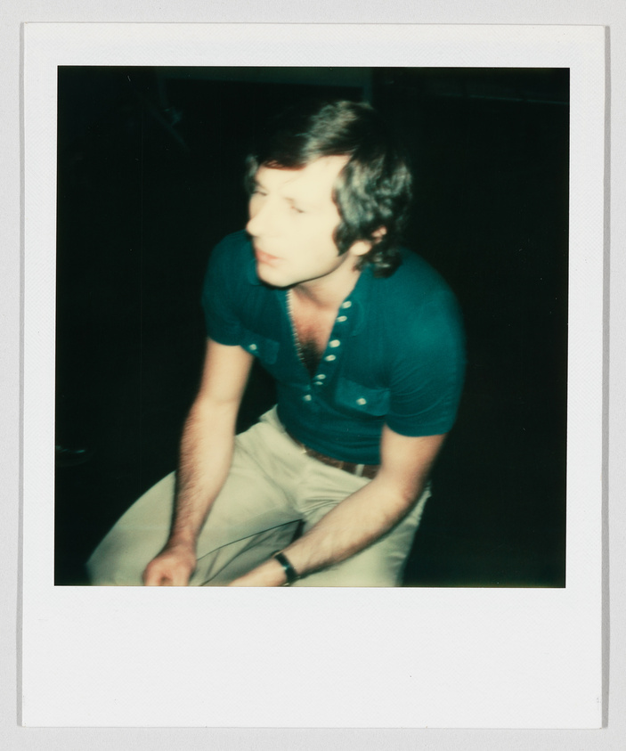 A man with short hair and a teal shirt sits, looking slightly downward, in a dimly lit space.