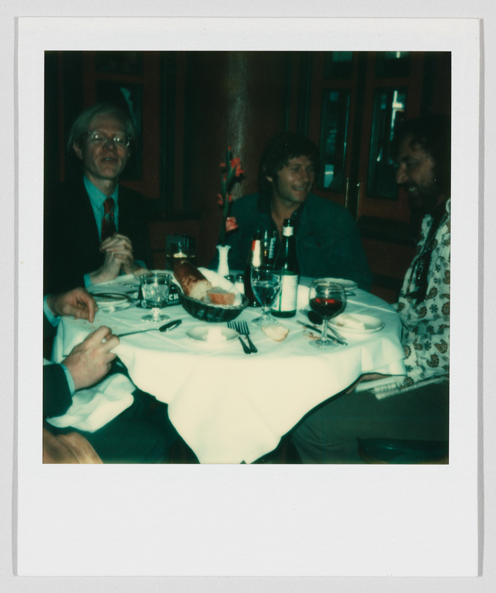 Three people sit around a restaurant table with drinks, bread, and wine, talking and smiling together.