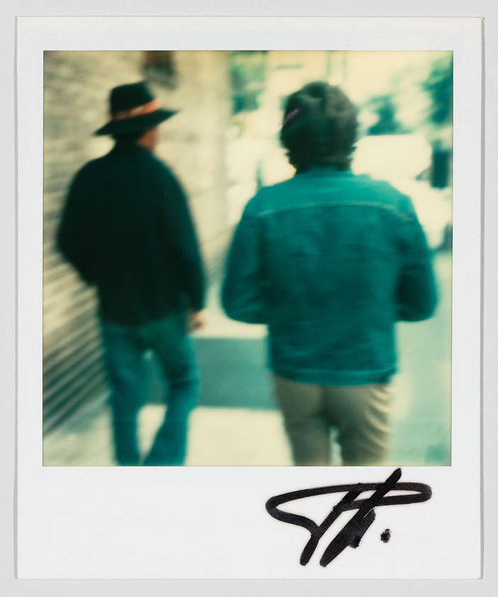 Two people walk down a sidewalk; one wears a wide-brimmed hat. A large signature is at the bottom.