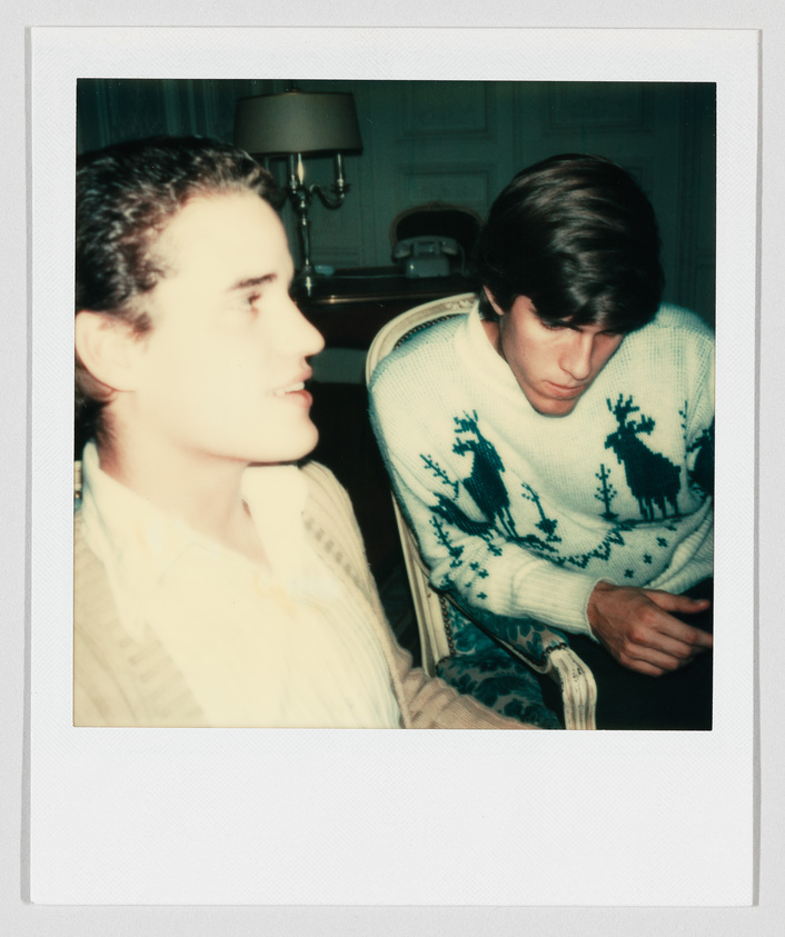 Two young men sit indoors; one wears a white sweater with blue reindeer patterns and looks down.