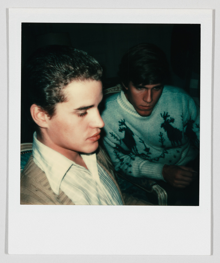 Two young men sit close together indoors, one wearing a striped shirt and the other a sweater with reindeer.