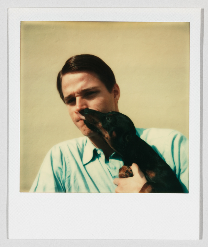 A man in a light blue shirt holds a small black dog that is licking his face.