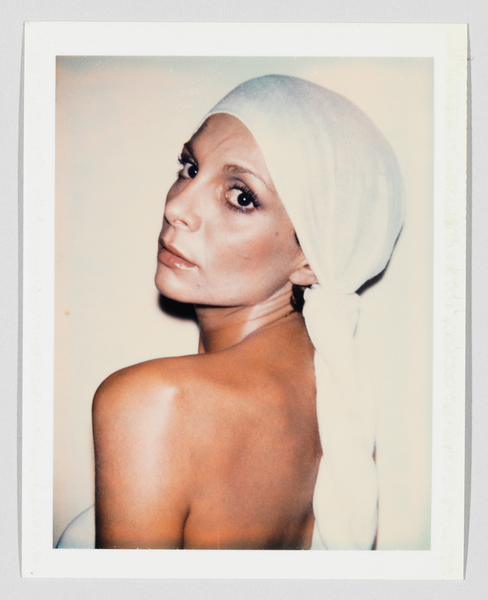 A woman with bare shoulders looks over her shoulder, wearing a white headscarf tied at the back.