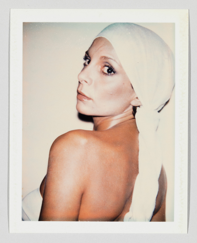 A woman with bare shoulders looks over her shoulder, wearing a white headscarf tied at the back.