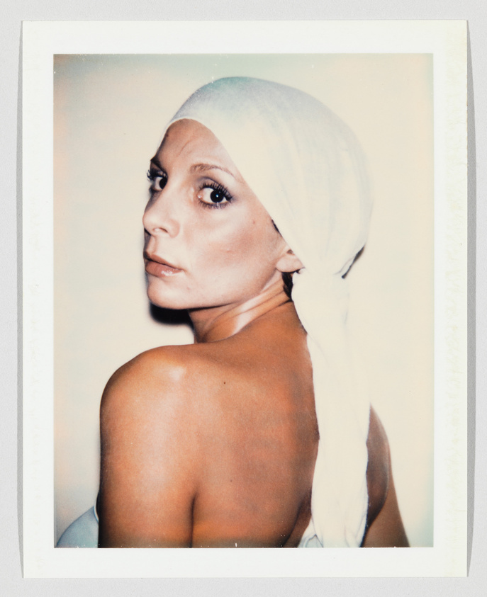 A woman with bare shoulders looks over her shoulder, wearing a white headscarf tied at the back.