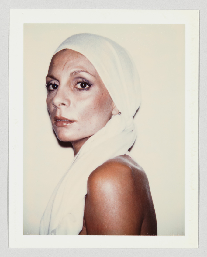 A woman with a serious expression wears a white headscarf, looking over her shoulder against a plain background.