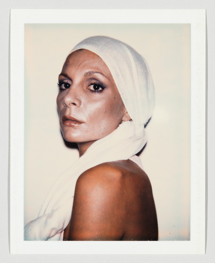 A woman with bare shoulders looks at the camera, wearing a white headscarf wrapped around her head.