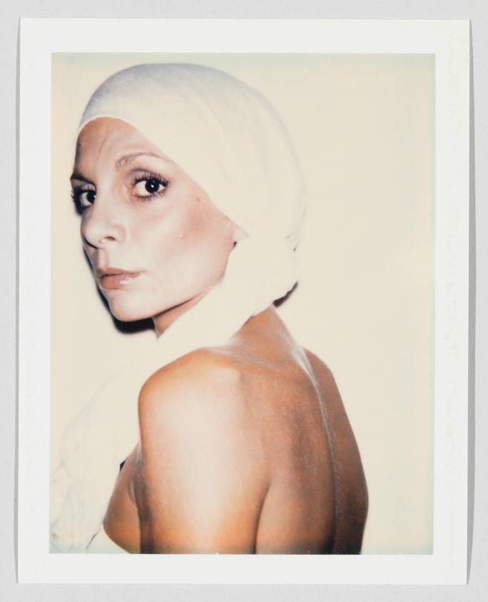 A woman with bare shoulders looks over her shoulder, wearing a white headscarf tied at the back.