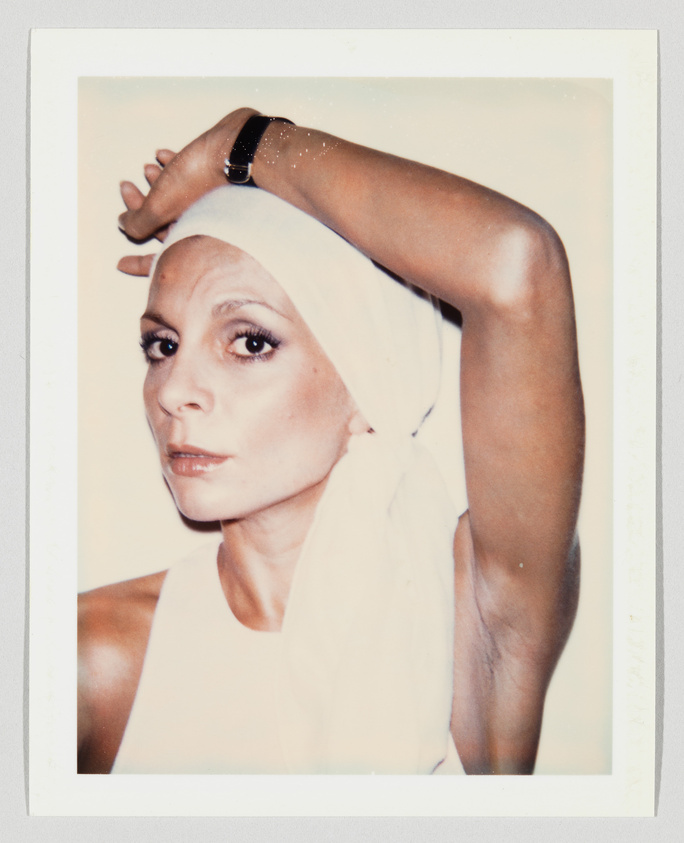 Woman wearing a white headscarf and sleeveless top, raising one arm above her head, looking at camera.