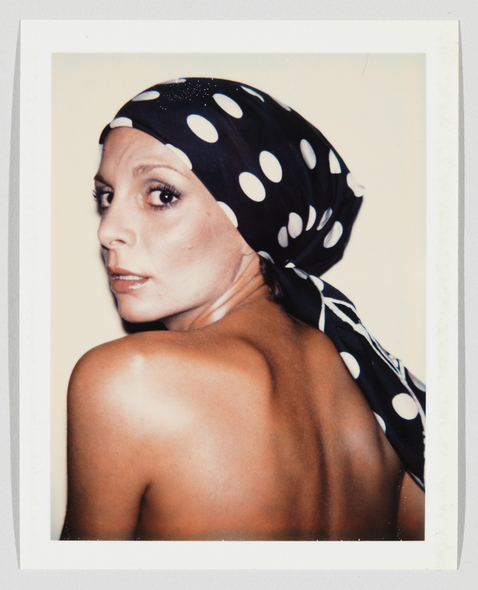 A woman with bare shoulders looks over her shoulder, wearing a black scarf with large white polka dots.