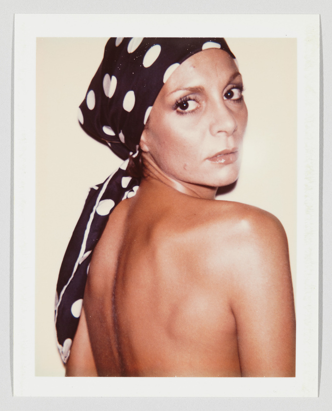 Woman with bare back and a large scar, wearing a black scarf with white polka dots, looking over shoulder.