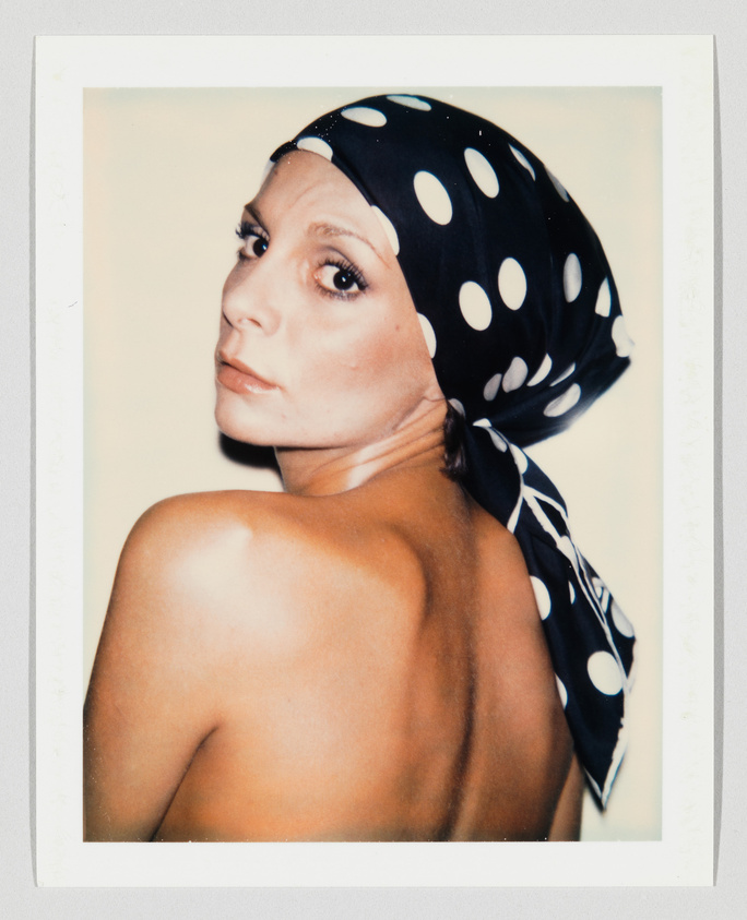 A woman with bare shoulders looks over her shoulder, wearing a black scarf with large white polka dots.