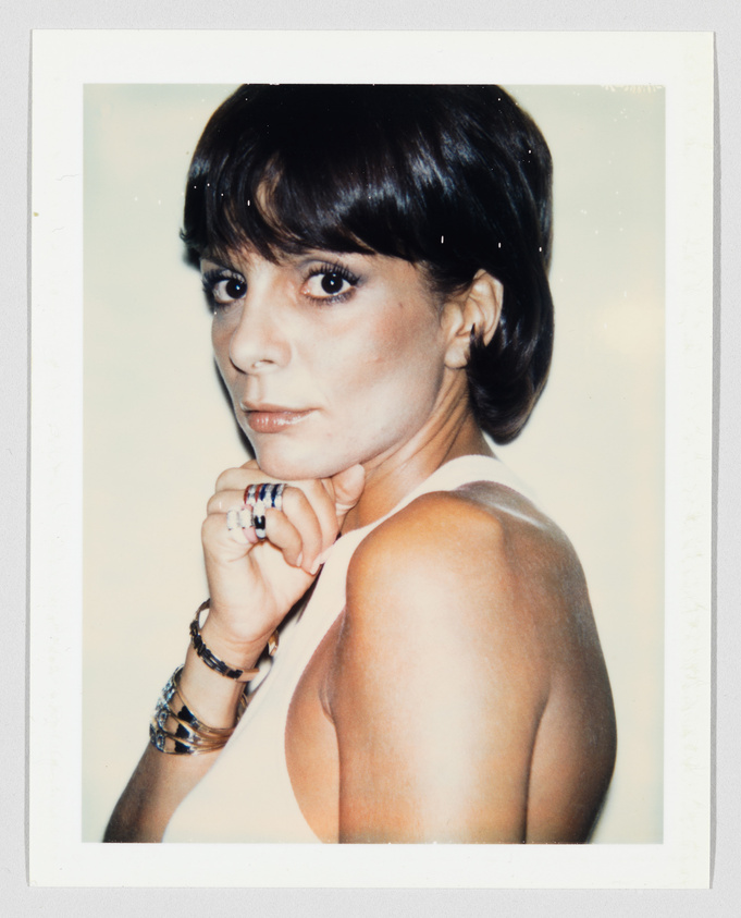 Woman with short dark hair looks over her shoulder, resting her chin on her hand, wearing bracelets.