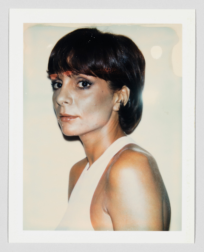 Woman with short dark hair and sleeveless white top looks over her shoulder with a neutral expression.