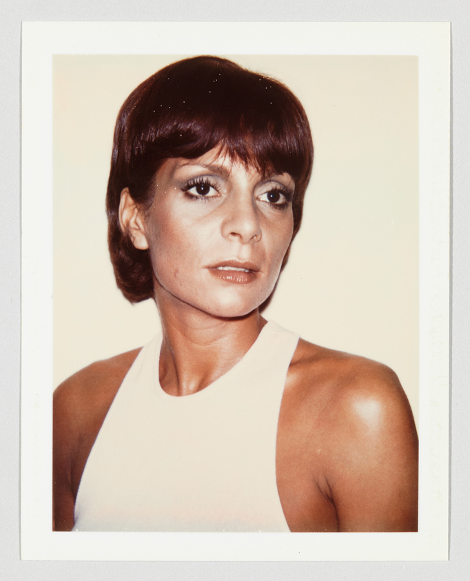 Person with short brown hair and a white sleeveless top looks slightly to the side with a neutral expression.