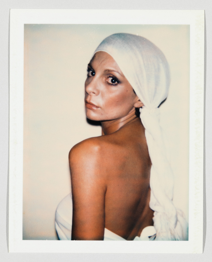 A woman with bare shoulders looks over her shoulder, wearing a white headscarf tied at the back.