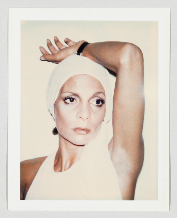 Woman wearing a white headscarf and sleeveless top, raising one arm above her head, looking serious.