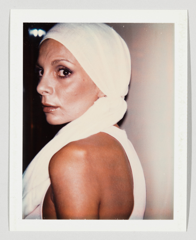 A woman with a white headscarf looks over her shoulder, facing the camera with a serious expression.