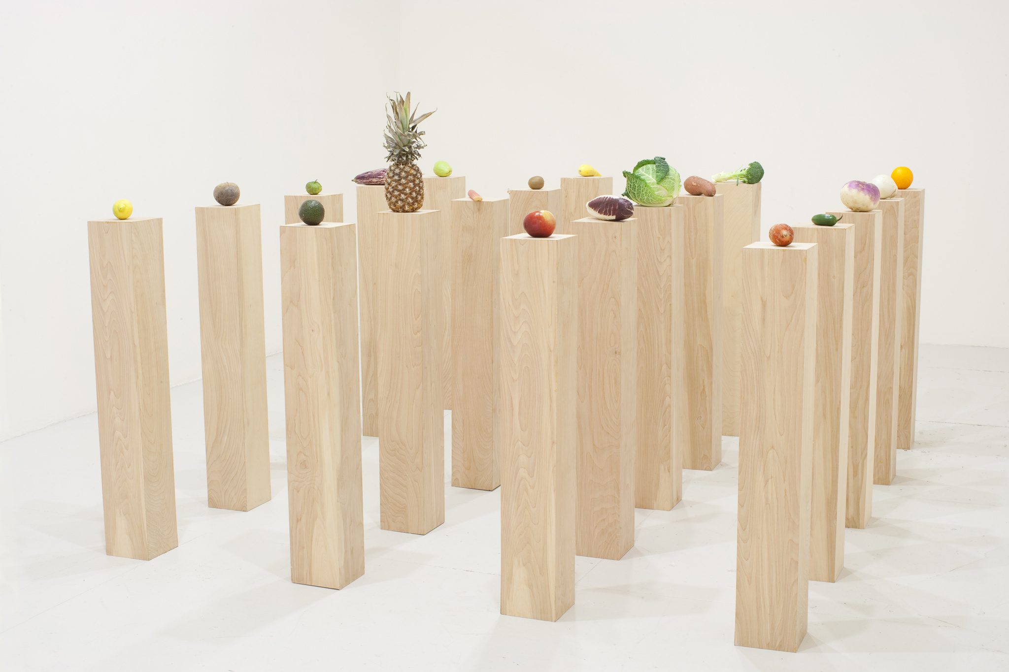 Wooden pedestals of varying heights with assorted fruits and vegetables on top in a white room.