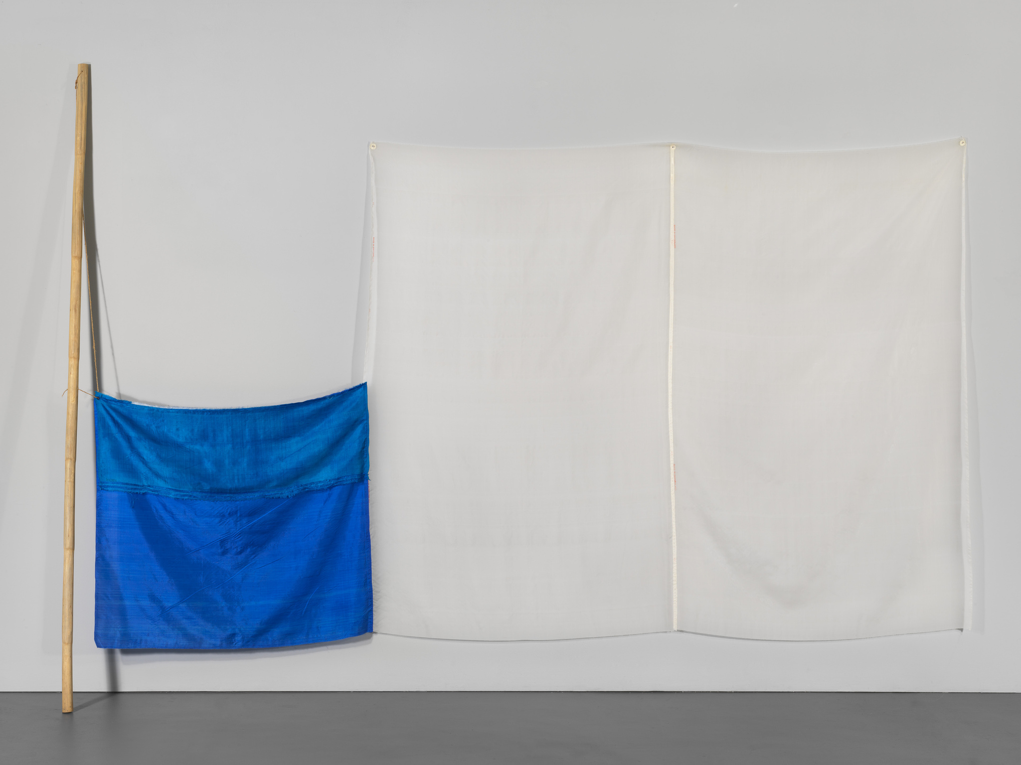 A blue fabric piece hangs from a wooden pole on the left, next to a larger white fabric piece suspended from the ceiling by two poles, against a grey wall.