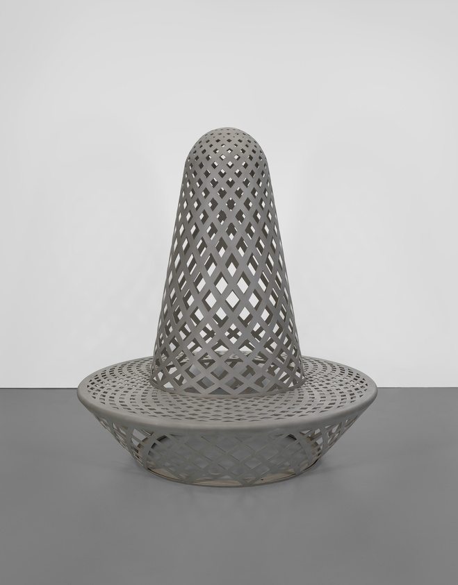 A large, gray, lattice-patterned sculpture with a conical top and circular base on a gray floor.