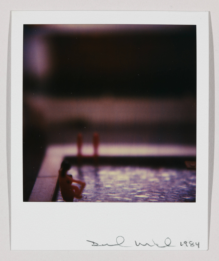 A Polaroid photo depicting a small, out-of-focus figurine sitting on the edge of a surface with a blurred background. The image has a vintage feel and includes a handwritten signature and the year 1984 at the bottom.