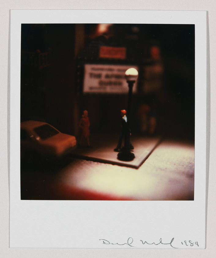 A vintage-looking photograph featuring a miniature scene with a street lamp illuminating a small area where two figurines, resembling a man and a woman, are standing near a car. The background is dimly lit, with a blurred sign that reads "THE BAND'S HERE." The image has a warm, reddish tone and is framed with a white border that includes a handwritten signature and the year 1984 at the bottom.