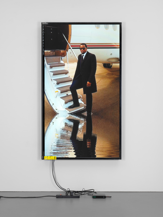 A man in a suit descends a private jet's stairs, reflected in a puddle. The image is displayed on a screen with visible cables below.