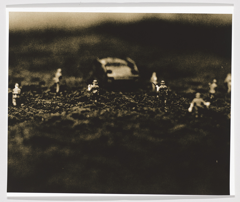 A sepia-toned photograph depicting a miniature scene with small figurines resembling soldiers scattered across a rough, uneven terrain. In the background, there is a blurred object that appears to be a vehicle. The image has a vintage or aged appearance, with a vignette effect darkening the corners.