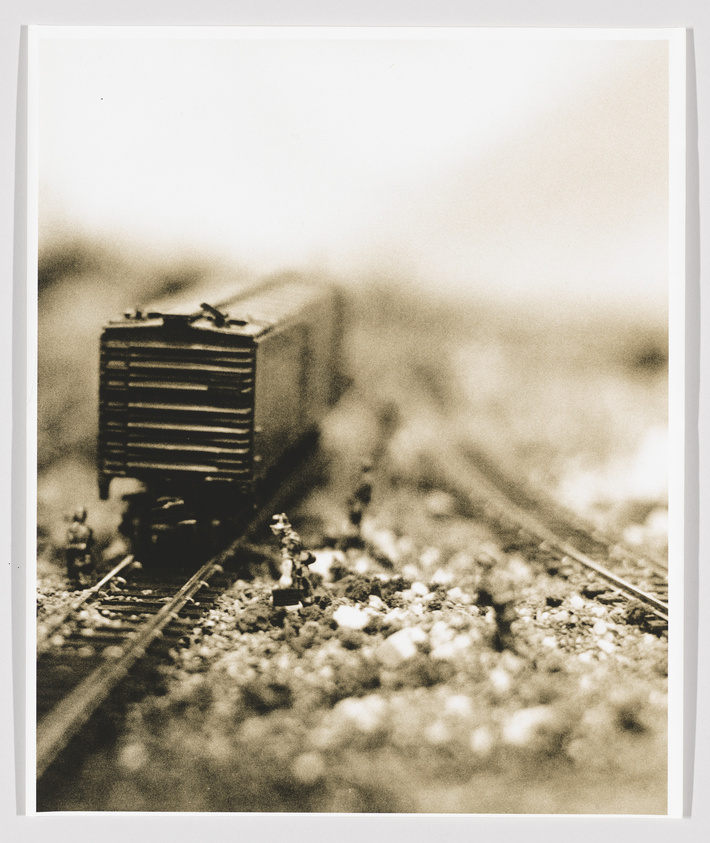 A sepia-toned photograph of a model train on tracks, with a shallow depth of field focusing on the front of the train, giving the scene a miniature, almost dreamlike quality.