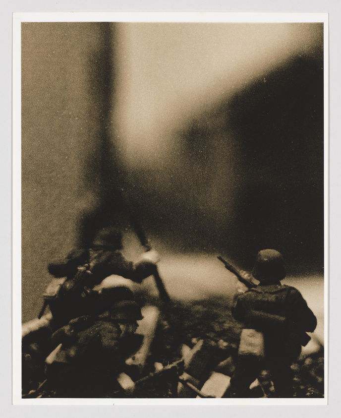Sepia-toned photograph depicting toy soldiers amidst rubble, with one figure in the foreground aiming a rifle, evoking a war scene. The image has a grainy, vintage appearance with soft focus and blurred background elements.