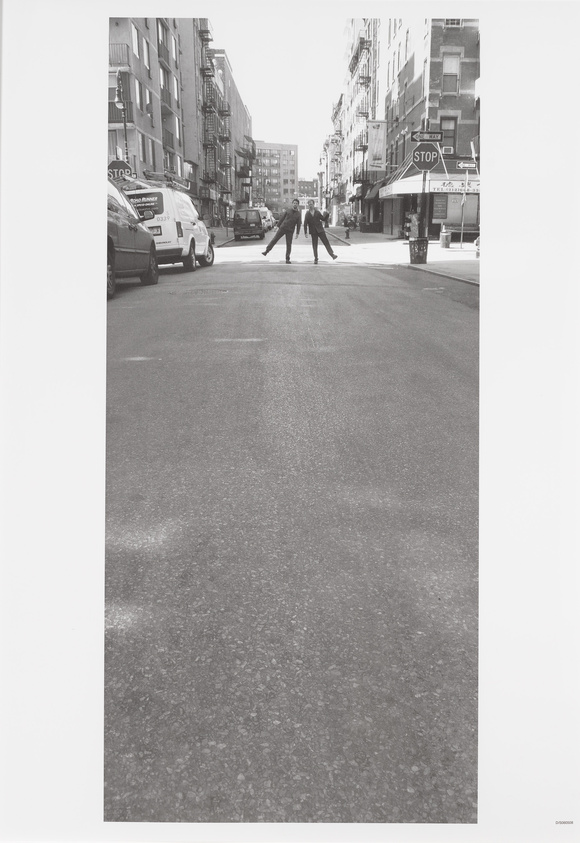 A black and white photograph capturing two individuals standing in the middle of a city street, arms outstretched towards each other, creating a mirrored pose. The street is lined with parked cars and buildings, and there's a stop sign and one-way sign visible on the right. The scene is devoid of traffic, giving a sense of stillness to the urban environment.