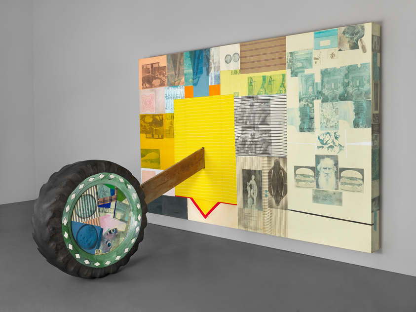 A large, colorful collage artwork is displayed on a gallery wall, featuring a variety of images and patterns in different textures and hues. In the foreground, a sculptural element resembling a large black tire with a mirrored center leans against the wall, with a wooden beam extending from its center towards the viewer. The reflection in the tire's mirror shows a distorted view of the collage and the gallery space.