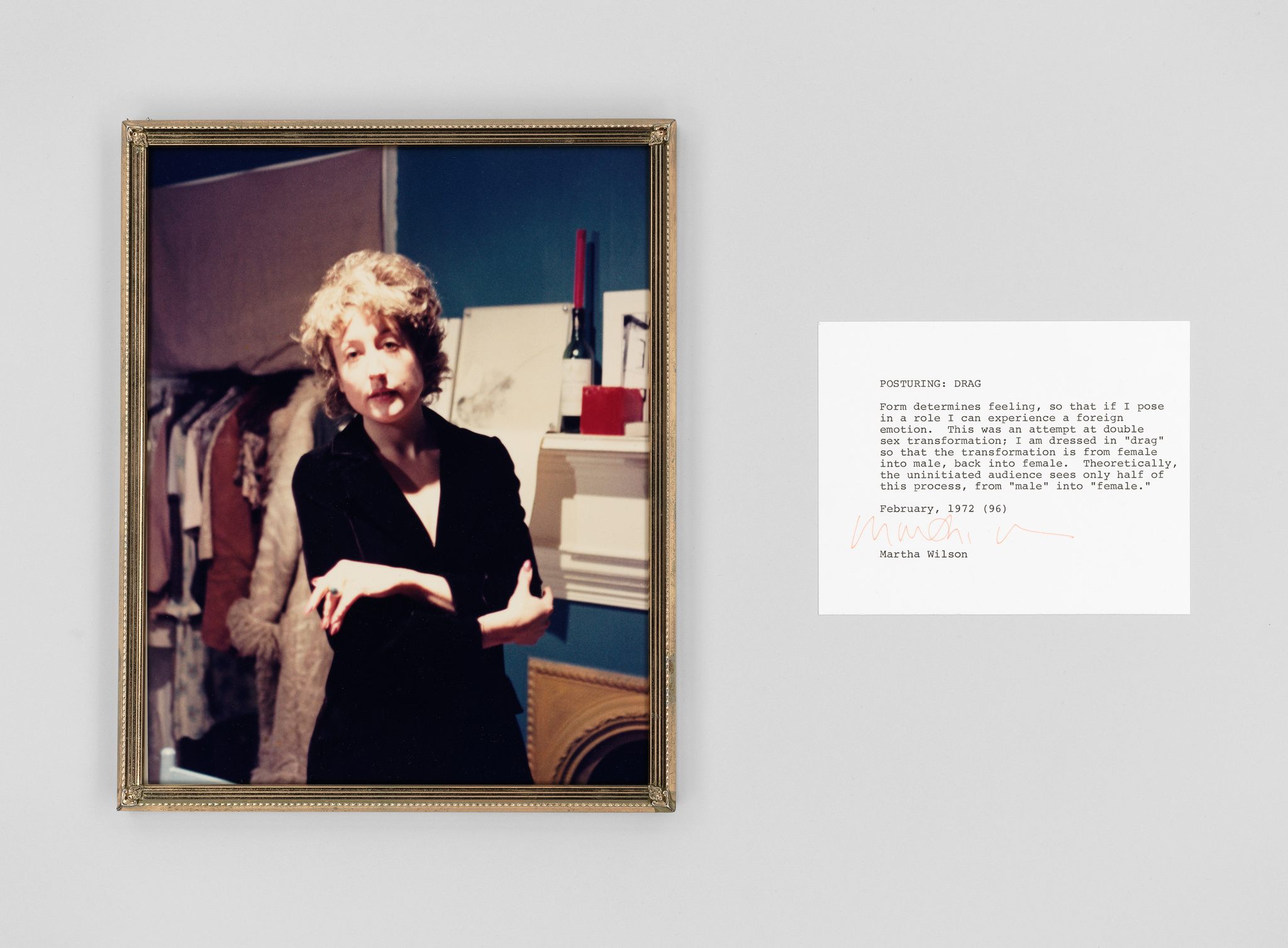 A framed photograph on a wall depicting a person with a mustache and short curly hair, dressed in a black blazer, posing with one arm crossed over their chest and the other resting on it. To the right of the photograph is a white card with typewritten text and a signature, explaining the concept of "posturing" in drag to express the fluidity of gender roles.