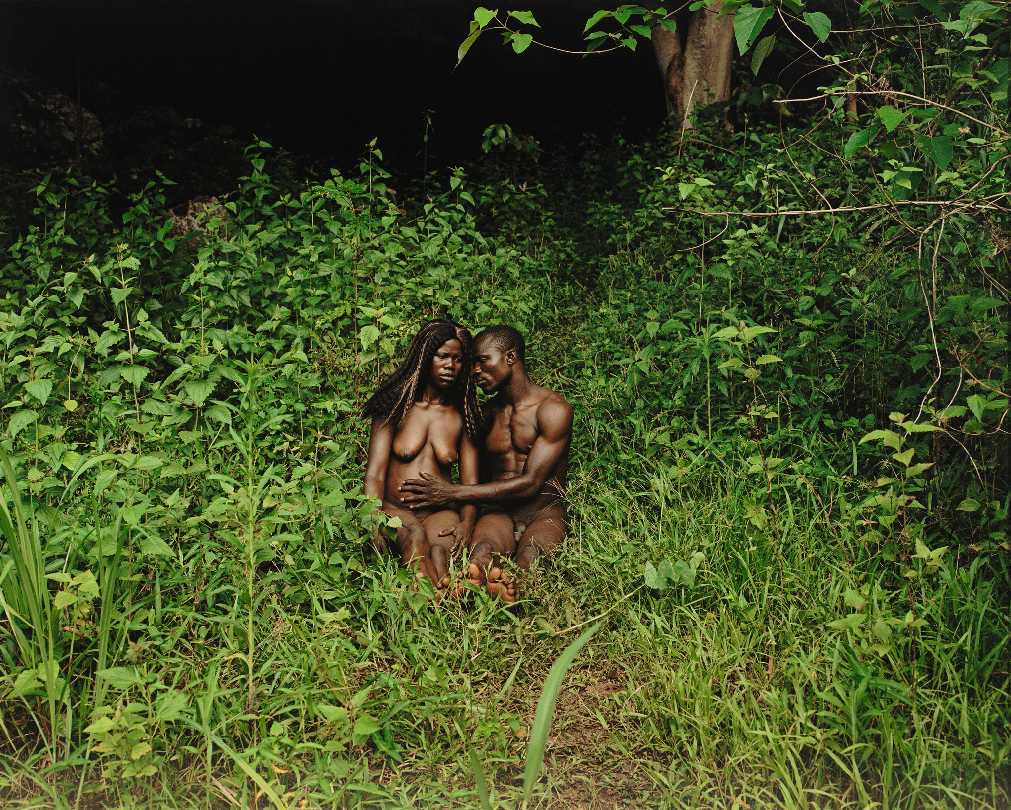 A Black couple sit in the nude in a lush green garden; the man's hand reaches across the woman's belly