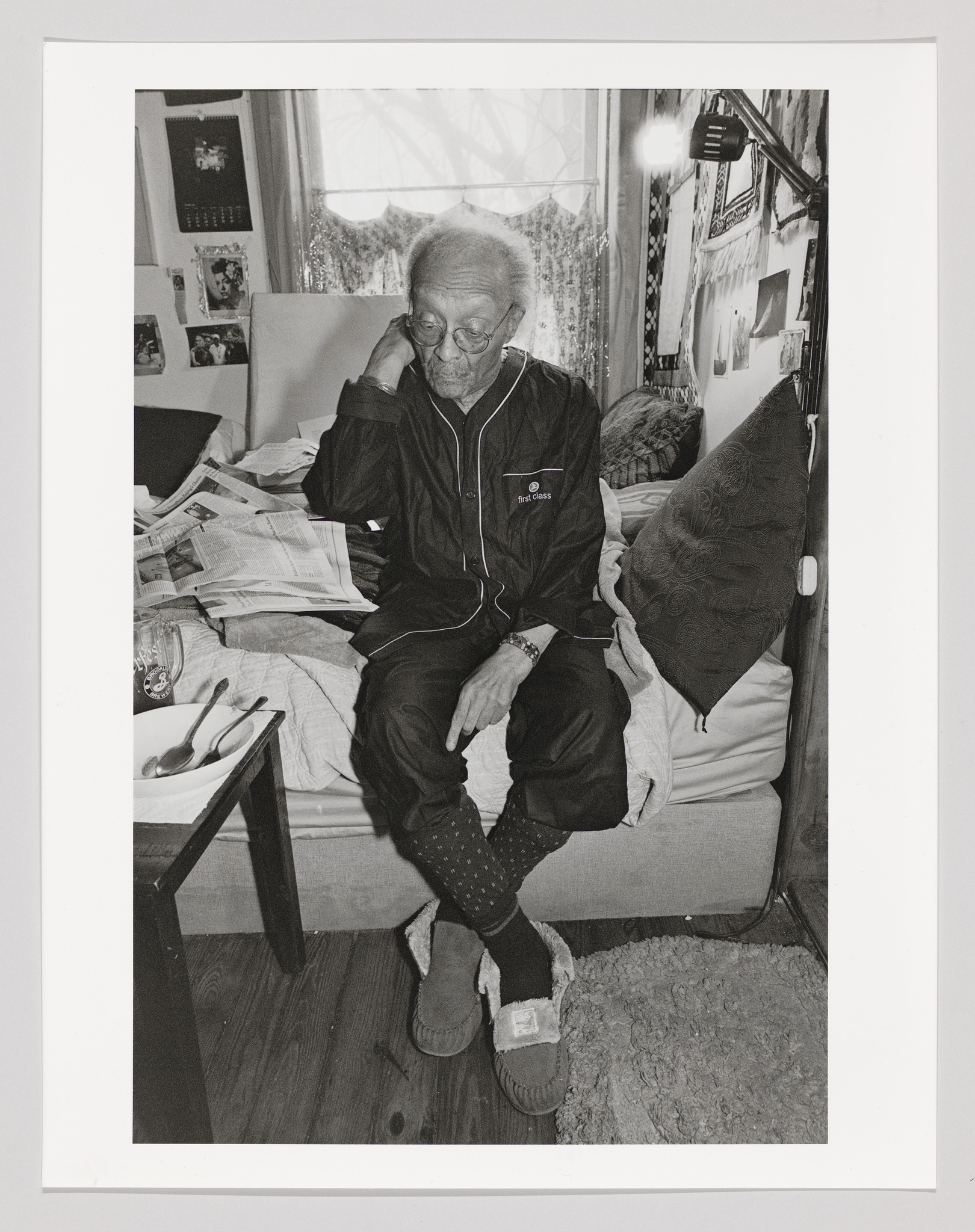 An elderly person with glasses is seated on a bed cluttered with newspapers, resting their head on their hand. They are wearing a black tracksuit with white stripes and polka-dot socks, and one slipper is on their foot while the other is on the floor. The room is cozy with personal items and photographs decorating the walls, and a window with curtains is partially visible in the background.