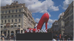 A large red high-heeled shoe sculpture is displayed in an urban square with classic European architecture and a partly cloudy sky in the background. The words "we look" are superimposed at the bottom of the image.