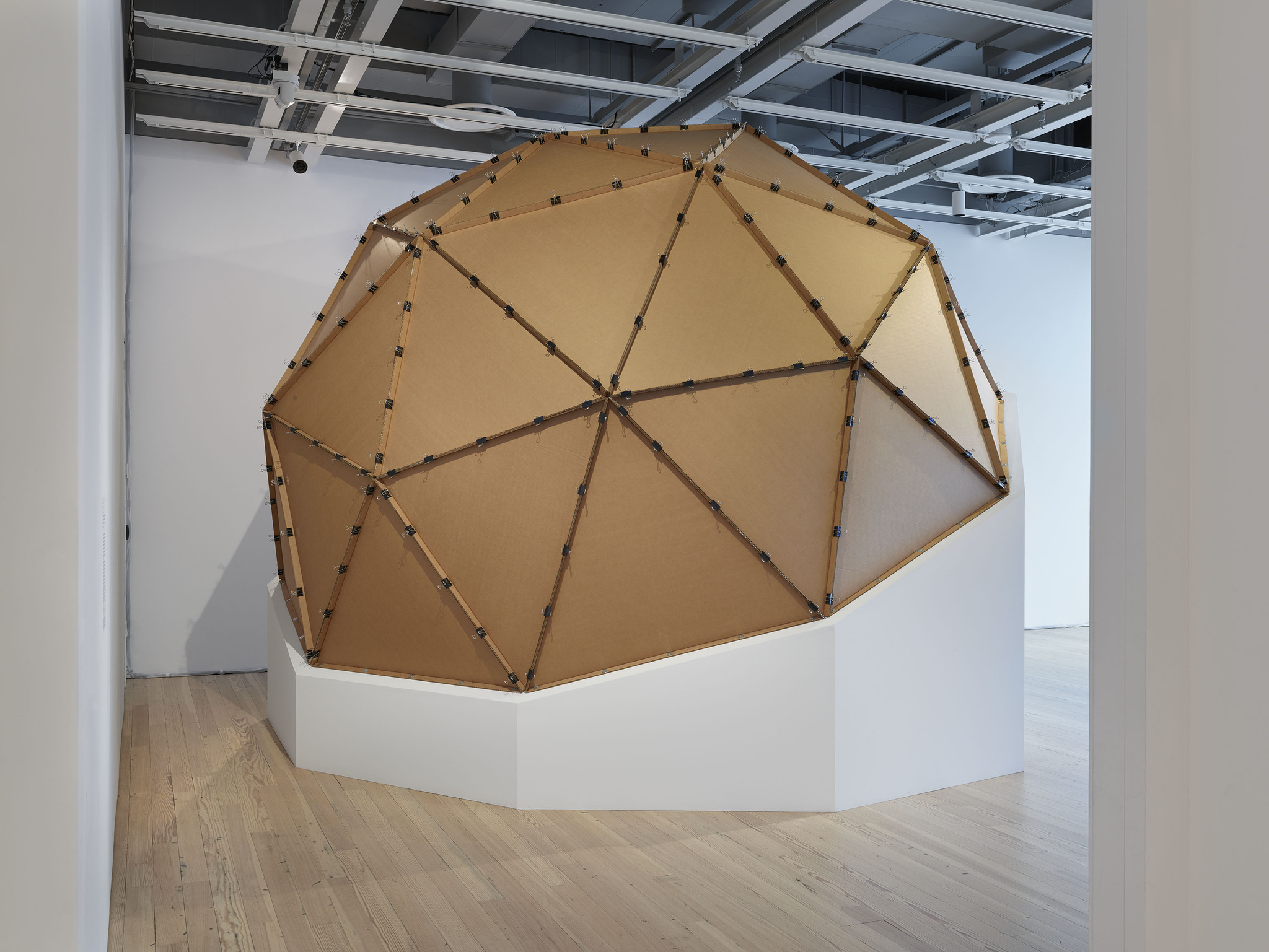 A large, geodesic dome-like structure made of cardboard panels connected by metal joints, displayed in a white-walled gallery space with wooden flooring and industrial ceiling fixtures.
