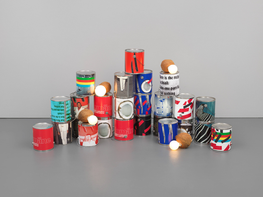 A collection of colorful and uniquely designed tin cans of various sizes arranged on a grey surface against a grey background. Some cans feature graphic patterns, others have images of people, and a few have text. Several cans are topped with coconuts, and one has a light bulb placed on it. The overall arrangement gives the impression of a modern art installation.