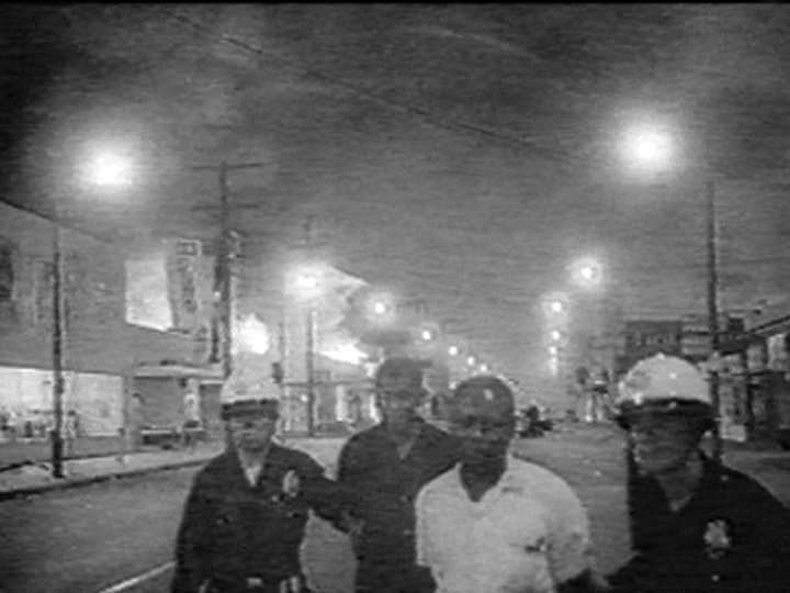 Three police officers escort a man down a city street at night, with smoke and fires visible in the background.