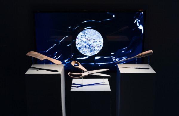 A modern art installation featuring three pedestals in front of a large monitor displaying a dynamic blue and white abstract image. Each pedestal holds an oversized common object: a comb, a pair of scissors, and a toothbrush, all with exaggerated proportions and positioned as if interacting with the swirling patterns on the screen.