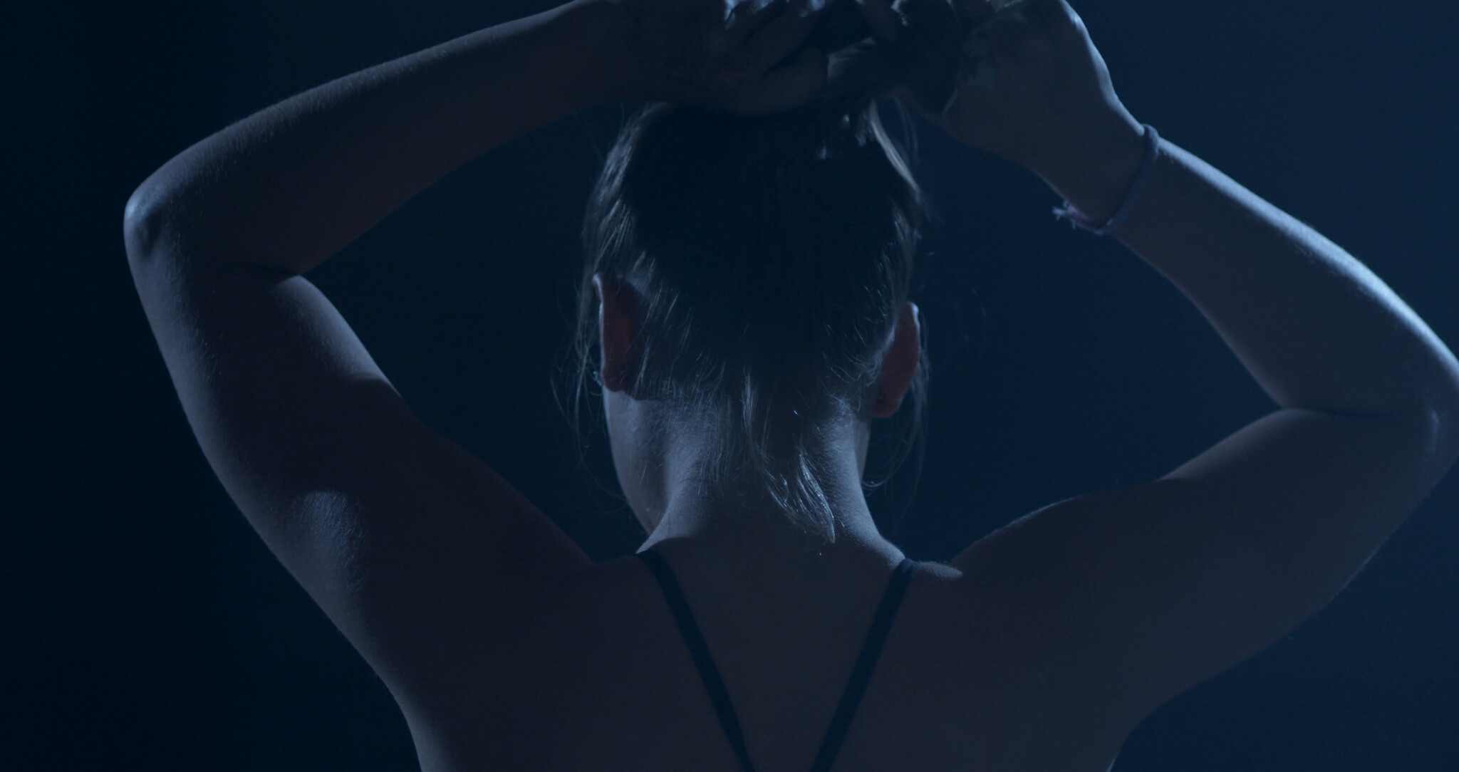 A silhouette of a person from behind, tying their hair up in a dark, moody setting. The individual's bare shoulders and arms are visible, and they are wearing a thin-strapped top. The lighting accentuates the contours of their arms and back.