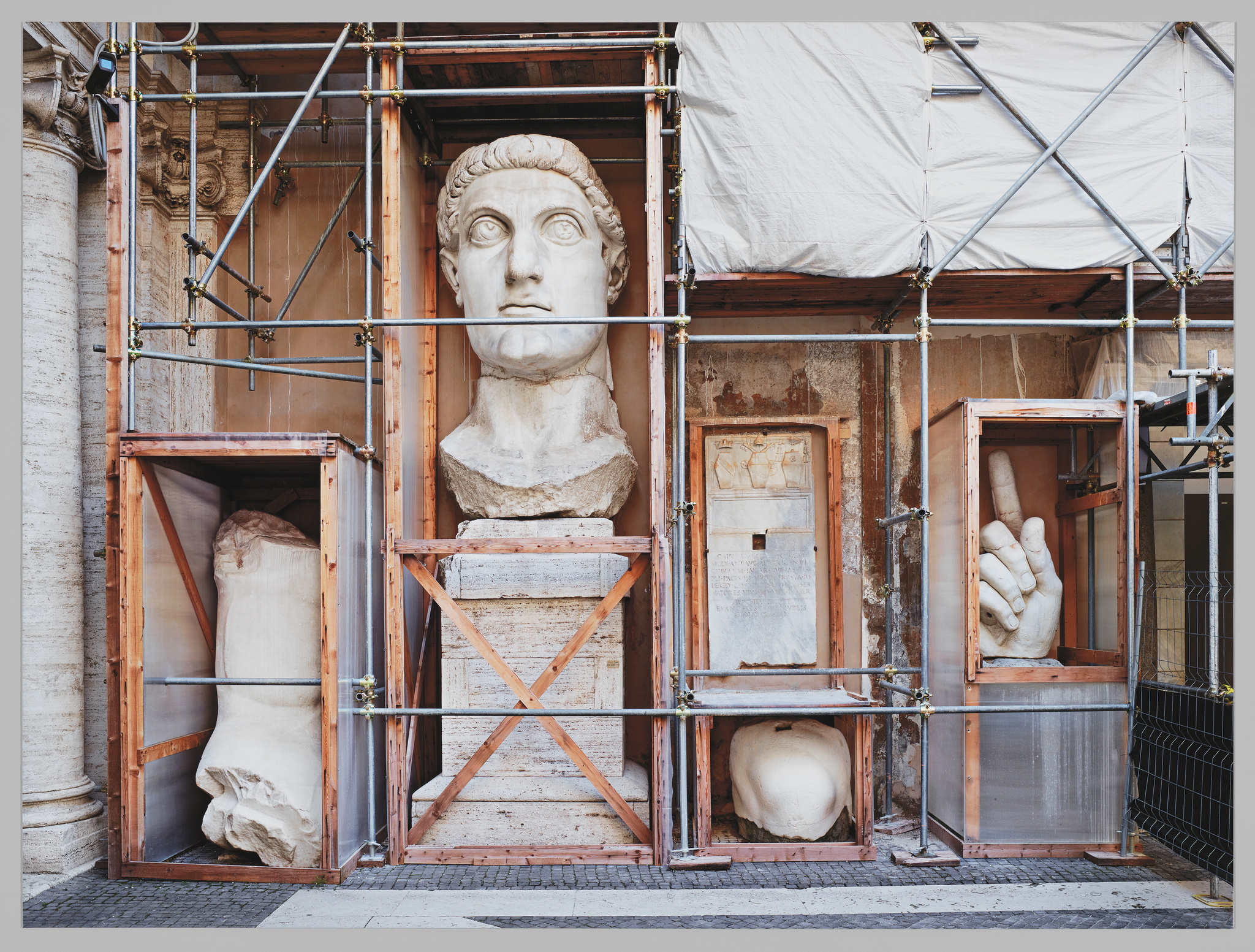 Giant statue parts in wooden crates during restoration, with a head, hand, and foot visible amidst scaffolding.