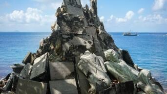 A pile of broken gravestones and statues stacked by the sea, with a boat visible in the distance.
