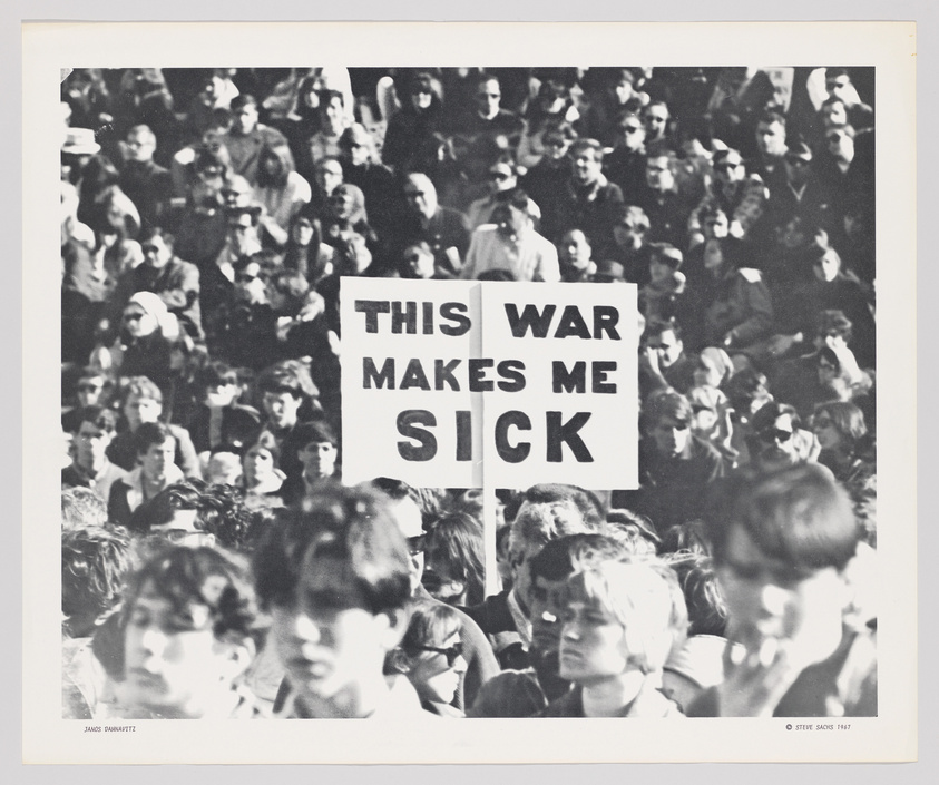 A black and white photograph capturing a dense crowd of people at a protest with a prominent sign held up that reads "THIS WAR MAKES ME SICK." The faces of the individuals are mostly indistinct, conveying the collective nature of the event. The photo is credited to Janos Panitz and Steve Sachs, 1987.