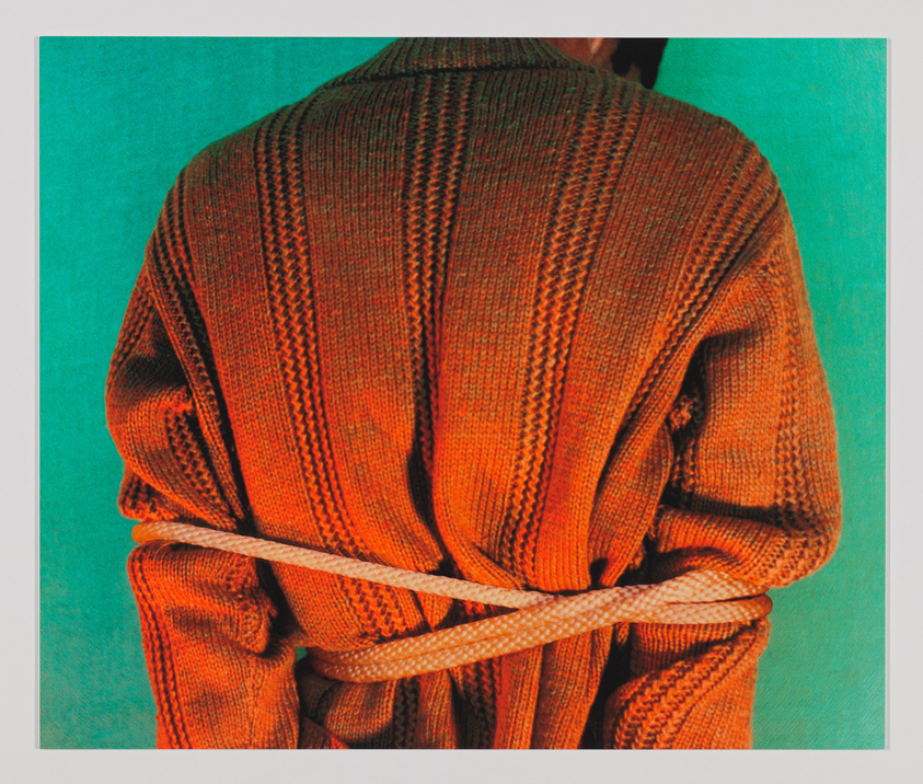 A person is seen from behind, wearing a textured orange sweater that is cinched at the waist with a braided brown belt. The background is a solid teal color, providing a stark contrast to the warm tones of the sweater.
