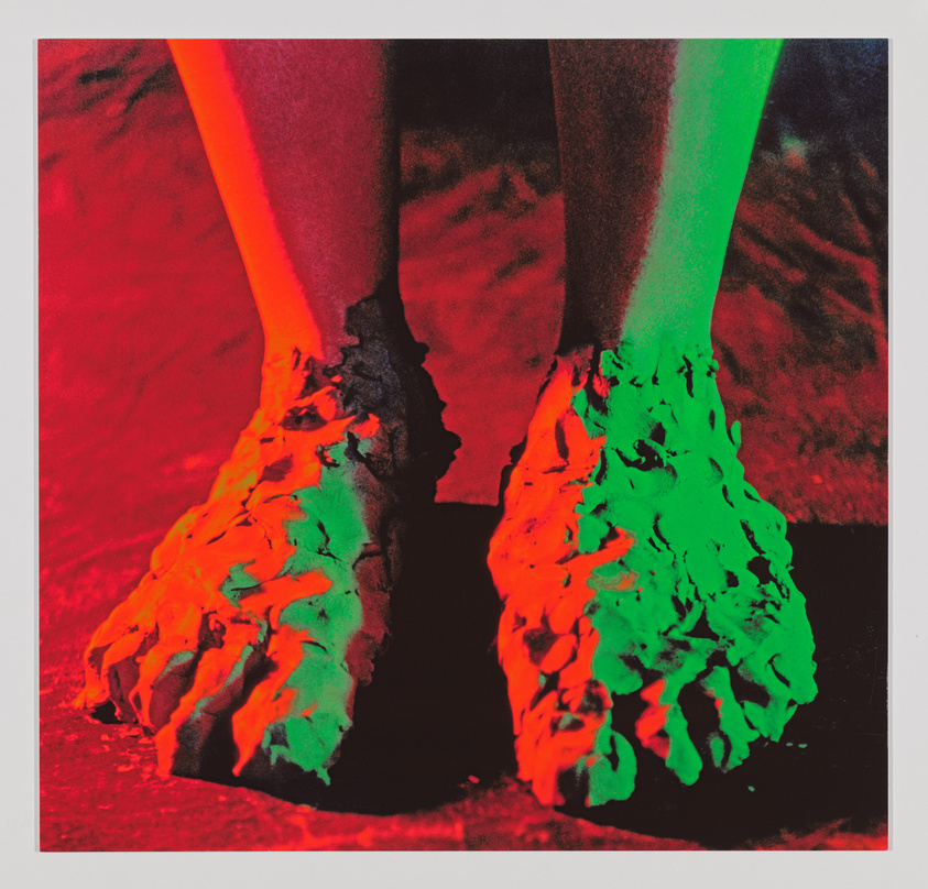A vividly colored photograph showing a pair of human feet covered in a thick, dripping substance, with one foot illuminated in red and the other in green, against a dark background with red hues.