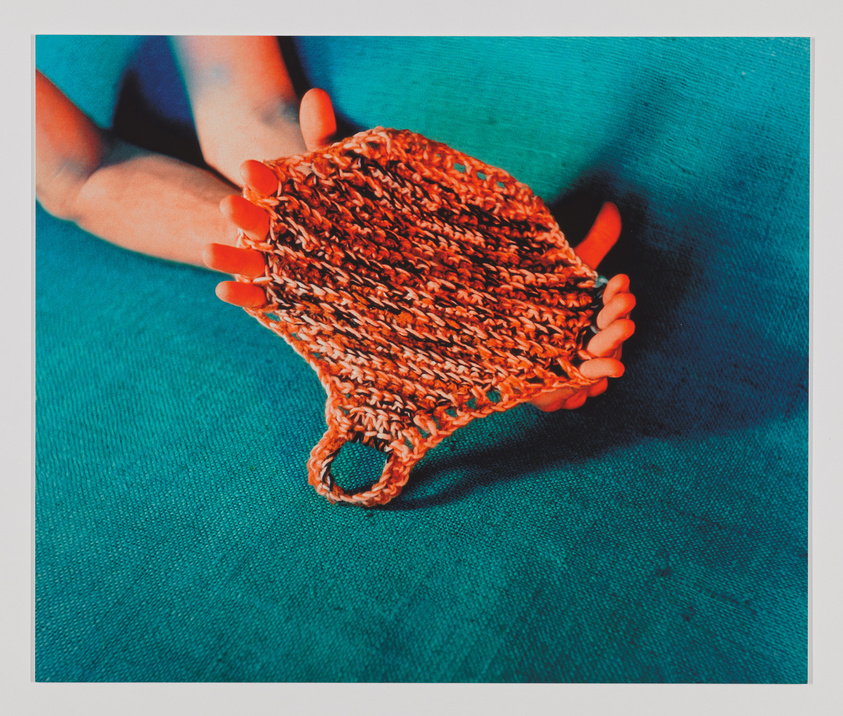 A close-up of a person's hands with bright orange nails holding a small, knitted orange and beige pouch against a teal fabric background.