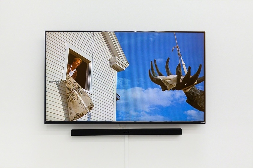 A flat-screen television mounted on a white wall displaying a scene with a person smiling and looking out of a window on the left, and a large moose head sculpture hanging by a rope on the right against a clear blue sky background.